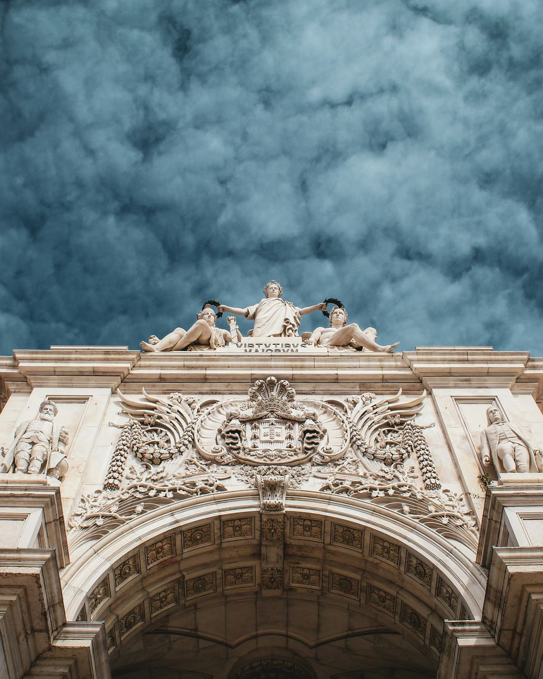 The carved top of the Rua Augusta Arch in Lisbon under a dark, heavy sky